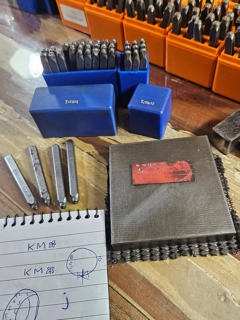 A collection of metal stamping dies organized in blue and orange plastic cases on a wooden workbench, alongside a hammer, a metal anvil block with a red sticker, and a handwritten notebook page.